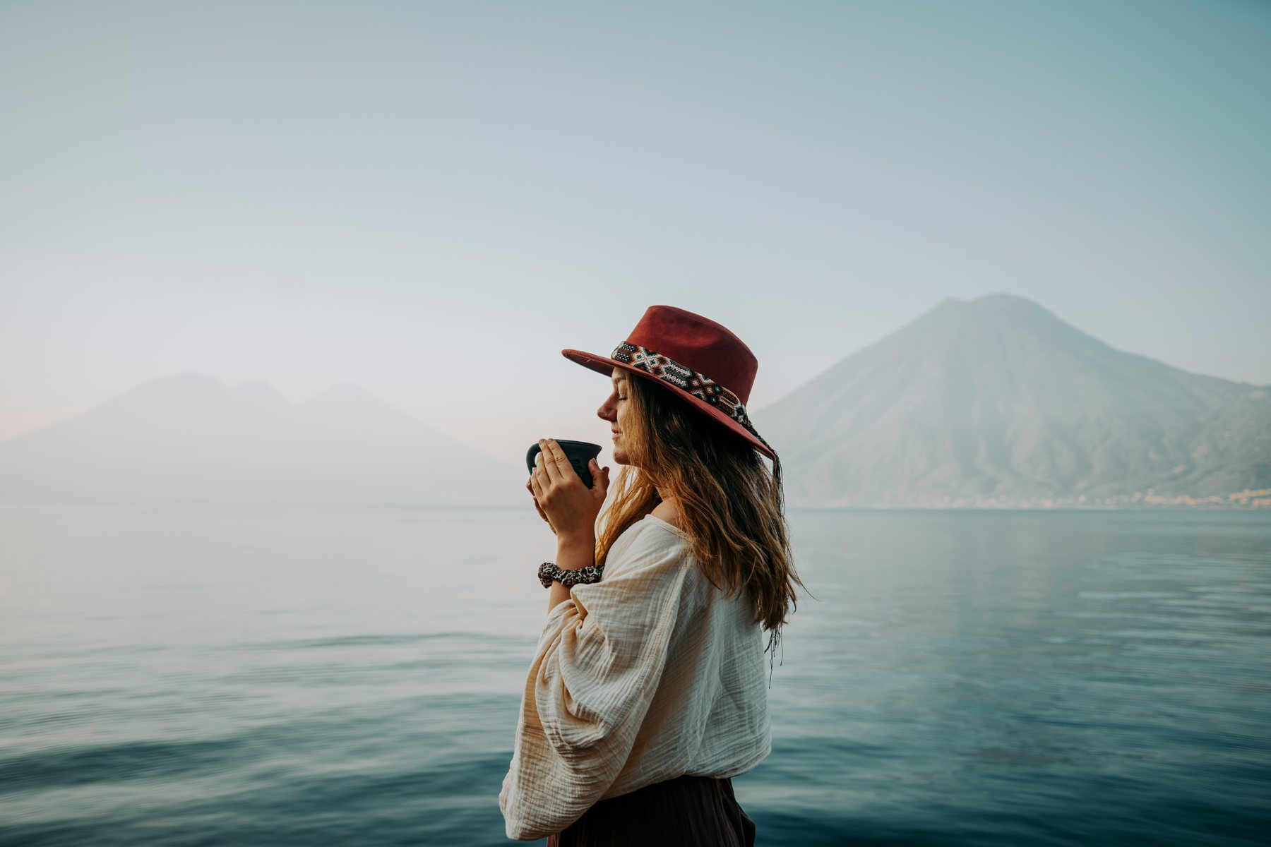 A woman savoring a warm cup of cacao by Lake Atitlán, eyes closed, a volcano behind her